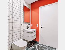 Modern bathroom with a wall-hung toilet and floating white vanity sink beneath a rectangular mirror, set against white grid wall tiles and a bold red accent wall. Dark marble-patterned floor tiles and a white door with black hardware complete the space.