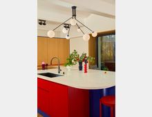 Moderne Küche mit einer Insel aus leuchtend roten und blauen Farbakzenten. Die Arbeitsplatte ist weiß mit einer eingebauten Spüle, dekoriert mit Vasen und Pflanzen; darüber hängt eine auffällige, minimalistische Deckenleuchte mit runden Leuchtkörpern.
