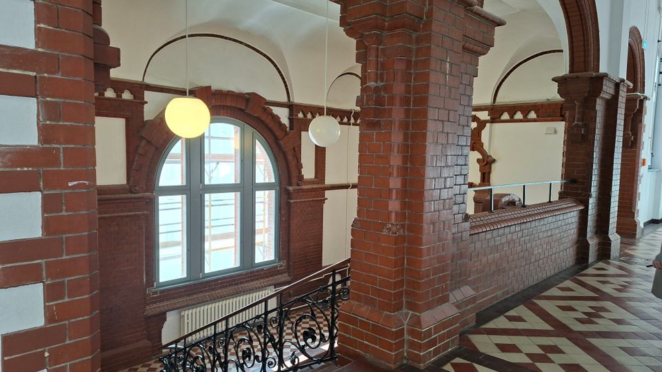Innenansicht eines historischen Flurs mit roten Backsteinpfeilern und Rundbögen, einer großen bogenförmigen Fensteröffnung und hängenden Kugelleuchten. Ein gemusterter Fliesenboden und ein schmiedeeisernes Geländer säumen die Galerie entlang der Wand.