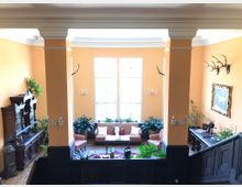 Heller Salon mit orangefarbenen Wänden und großen Fenstern, ausgestattet mit antiken Holzmöbeln, einem Sofabereich mit gemusterten Polstern, Pflanzen und Geweih-Dekorationen an den Wänden. Rechts führt eine dunkle Holztreppe hinunter, und ein gerahmtes Gemälde hängt an der Wand.