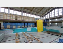 Ein leerstehendes Hallenbad mit leeren Sitzreihen im trockenen Becken, bröckelnden Wänden und einer auffälligen gelben Stoffbahn, die von der oberen Galerie hängt. Die Umgebung zeigt alte Umkleidekabinen über zwei Ebenen und große Fenster, die natürliches Licht einlassen.