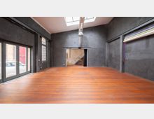 Großer, leerer Raum mit dunklen Wänden und einem Holzboden, einer Glas-Doppeltür auf der linken Seite, hohen Fenstern und einem Dachfenster, das Tageslicht einlässt. Im hinteren Bereich führen Stufen zur höheren Ebene, und an der rechten Wand befinden sich aufgerollte bunte Stoffbahnen.