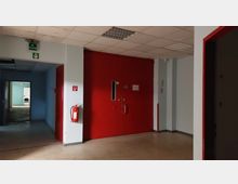 Ein Flur in einem Gebäude mit weißen Wänden und roten Türen, darunter eine große zweiflügelige Tür mit kleinen Fenstern. An der Wand befindet sich ein Feuerlöscher und ein Fluchtweg-Schild, der Boden ist mit hellem Linoleum ausgelegt.