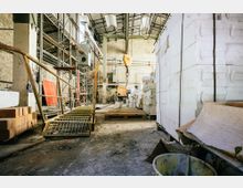 Industrielle Lagerhalle mit Metallgerüsten, gestapelten weißen Blöcken und Baumaterialien wie Ziegeln und Plastikeimern; im Hintergrund sind ein Deckenkran und Stahlträger sichtbar.