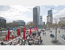 Ein städtischer Platz mit Restaurants und Cafés im Vordergrund, deren Tische und rote Sonnenschirme gut sichtbar sind. Im Hintergrund stehen hohe moderne Gebäude und ein Baukran, umgeben von Fußgängern und Radfahrern.