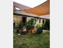 Ein ländlicher Hof mit einer Ziegelsteinfassade und roten Dachziegeln. Im Vordergrund stehen mehrere Töpfe mit Pflanzen und Blumen, daneben eine Schubkarre, während ein Sonnensegel über dem Bereich gespannt ist.