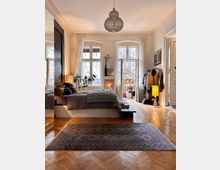 Helles Schlafzimmer mit Holzboden und großem Teppich, ausgestattet mit einem Bett, Pflanzen und Deko-Elementen. Im Hintergrund befinden sich hohe Fenster mit Vorhängen, ein kleiner Kamin und ein Zugang zu einem Balkon.