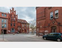 Straßenszene mit historischen Backsteingebäuden in gotischem Stil mit spitzbogigen Fenstern und verzierten Fassaden. Im Vordergrund stehen ein Baum, ein geparktes Auto und ein Straßenlaterne, während im Hintergrund ein kleiner gepflasterter Platz zu sehen ist.