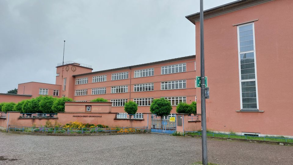 Backsteinfarbenes Schulgebäude mit flachem Dach, großen Fensterfronten und einem zentralen Turm mit Uhr. Vor dem Gebäude befinden sich ein Metalltor, eine gepflasterte Fläche und gepflegte Grünanlagen mit Blumenbeeten und kleinen Bäumen.