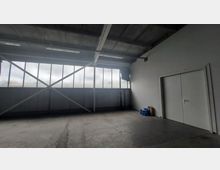 Ein leerer, industriell wirkender Raum mit Betonboden, einer Deckenstruktur aus Metallträgern und großen Fenstern mit Milchglas, die teilweise durch Metallkreuze verstärkt sind. Rechts befindet sich eine weiße Tür vor einer glatten Wand, und es liegen einige Gegenstände, darunter eine blaue Tasche, auf dem Boden.