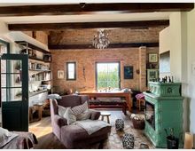 Wohnzimmer mit gemütlicher Einrichtung in rustikalem Stil. Sichtbare Ziegelwände, Holzbalken an der Decke, ein grüner Kachelofen, ein großer Holztisch vor einem Fenster sowie ein Sessel und offene Regale prägen den Raum.