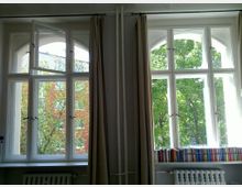 Zwei große, weiße Fenster mit Bogenoberseiten in einem Innenraum, durch die Bäume und eine rote Gebäudeaußenwand sichtbar sind. Vor den Fenstern stehen Heizkörper, und auf der Fensterbank sind Bücher in verschiedenen Farben zu sehen; beidseitig hängen beige Vorhänge.