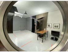 Ein moderner Raum mit grauen Wänden und einem hellen Boden, durch eine runde Glasscheibe fotografiert. Zu sehen sind ein Holzarbeitstisch, Fotostudioausrüstung mit Beleuchtung, ein Monitor auf einem kleinen Tisch sowie zwei gerahmte Bilder an der Wand.