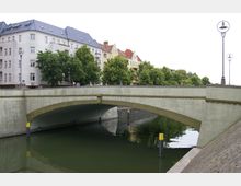 Steinbrücke über einen Kanal in einer städtischen Umgebung mit Wohnhäusern im Hintergrund. Die Brücke hat ein schlichtes Design mit einer Verzierungsmaske im Zentrum, und der Kanal ist von Bäumen und Straßenlaternen gesäumt.