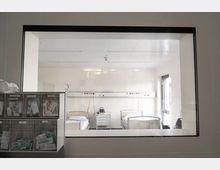 Krankenhauszimmer mit zwei Krankenhausbetten, einem Nachttisch in der Mitte und medizinischer Ausstattung an der Wand. Im Vordergrund ist eine Sichtscheibe zu sehen, durch die der Raum betrachtet wird; rechts gelangt Tageslicht durch ein Fenster.