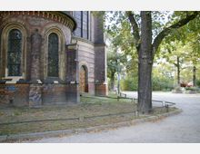 Teilausschnitt einer historischen Backsteinkirche mit Rundbogenfenstern und verzierten Steinstrukturen, umgeben von einem Park mit Bäumen und Fußwegen. Im Hintergrund sind ein Holztor und eine Straßenlaterne zu sehen.