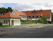 Historisches Gebäudeensemble mit roten Ziegeldächern, bestehend aus einem größeren Fachwerkgebäude im Hintergrund und einem kleineren, weiß verputzten Anbau im Vordergrund. Die Umgebung umfasst eine gepflasterte Straße, Rasenflächen und vereinzelte Bäume.