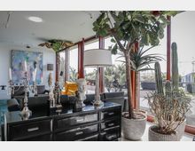 Helles Wohnzimmer mit großen Fensterfronten, moderner Einrichtung und zahlreichen Pflanzen, darunter Kakteen und ein großer Feigenbaum. Im Vordergrund ein schwarzes Sideboard mit dekorativen Skulpturen, Kerzenständern und einer Tischlampe.