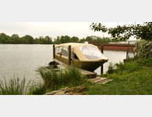 Boot mit beigem Verdeck, das an einem hölzernen Steg am Ufer eines Flusses befestigt ist. Im Hintergrund sind Bäume am gegenüberliegenden Ufer und ein Frachtschiff auf dem Wasser zu sehen.