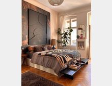 Ein gemütliches Schlafzimmer mit einem großen Bett, bedeckt von einer bunt gemusterten Decke, Holzfußboden und einer Wanddekoration mit floralen und abstrakten Elementen. Ein großes Fenster mit weißen Vorhängen lässt Tageslicht herein, daneben stehen Zimmerpflanzen und eine kleine weiße Kommode mit Dekorationen.