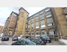 Mehrstöckiges Backsteingebäude mit großen Fenstern, kombiniert aus unverputztem Mauerwerk und teilweise grauem Putz, umgeben von einem Parkplatz mit geparkten Autos. Der Gebäudekomplex hat eine industrielle Bauweise mit einem markanten Turm und Arkaden über Türen und Fenstern.