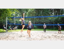Zwei Frauen spielen Beachvolleyball auf einem sandigen Spielfeld mit einem blauen Netz, umgeben von grünen Bäumen und Sträuchern. Im Hintergrund sitzen Zuschauer auf Stühlen im Gras.