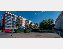 Wohnanlage mit fünfstöckigen Gebäuden, gekennzeichnet durch bunte Balkone in Blau und Gelb sowie Blumendekorationen. Im Vordergrund befinden sich ein gepflasterter Weg, Absperrpfosten und gepflegte Grünflächen mit Bäumen und Hecken.