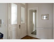 Helle Innenansicht eines Altbaus mit einer großen weißen Doppelflügeltür mit geätzten Glasscheiben, einem goldfarbenen Türgriff und Stuckverzierungen. Rechts daneben befindet sich eine offene Tür zu einem angrenzenden Raum und ein gerahmtes Bild an der Wand, der Boden ist mit Holzparkett ausgelegt.