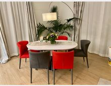 Ein runder Esstisch mit einer weißen Tischplatte, umgeben von Stühlen mit rotem und grauem Polster, steht auf einem Holzfußboden. Im Hintergrund befinden sich helle Vorhänge und eine Zimmerpflanze, beleuchtet von einer bogenförmigen Stehlampe.