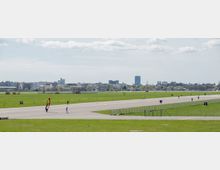 Weitläufiges ehemaliges Flugfeld mit einer breiten Asphaltlandebahn, umgeben von Grünflächen; vereinzelt sind Menschen zu Fuß oder mit Sportgeräten unterwegs. Im Hintergrund ist eine städtische Skyline mit niedrigeren Gebäuden und einem hohen Turm zu sehen.