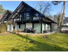 Zweistöckiges Wohnhaus mit schwarzem Holzgebälk und schrägen Dächern, großen Glasfronten sowie Balkonen im Obergeschoss. Im Vordergrund ein gepflegter Garten mit Rasen, Terrasse und einzelnen Gartenmöbeln, umgeben von Bäumen und Sträuchern.