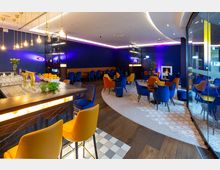 Moderne Lounge mit dunkelblauen Wänden und farbigen Polstersesseln in Blau und Orange, ergänzt durch runde Tische. Im Vordergrund ist eine Bar mit gelbem Beleuchtungselement und Hockern, während im Hintergrund lila Akzentbeleuchtung und ein eingebauter Kamin zu sehen sind.
