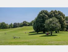 Weitläufige Parklandschaft mit großen Rasenflächen, sanften Hügeln und mehreren Bäumen, die Schatten spenden. Menschen sitzen oder liegen auf der Wiese, einige nutzen einfache Holzbänke oder Stühle.