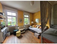 Wohnzimmer mit Holzdielenboden, gelb gestrichenen Wänden und großen Fenstern mit Blick ins Grüne. Der Raum ist möbliert mit einem grauen Sofa, bunten Kissen, einem Hochbett mit Vorhang sowie einem Regal und Sitzhockern.