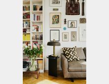 Wohnzimmer-Ecke mit einem grauen Ledersofa, dekoriert mit einem grafisch gemusterten Kissen, daneben ein moderner Beistelltisch mit einer gold-schwarzen Lampe. Die weiße Wand ist mit einer Vielzahl von Kunstwerken, darunter Gemälde und Textilien, dekoriert, und ein offenes Bücherregal ist voller Bücher und Dekorationsgegenstände.