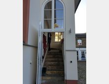 Eingang zu einem Haus mit offen stehender Tür, die eine schmale Treppe nach oben zeigt. Über der Tür ist ein großes Fenster mit halbrundem Abschluss, und im Hintergrund ist ein gepflasterter Hof sowie ein weiteres Gebäude sichtbar.