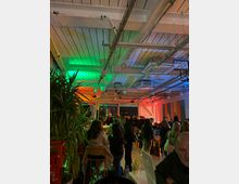 Interior of a modern event space with an exposed white-beam ceiling, visible ducts and cable trays, and colorful green, orange, and blue lighting. The room has tall plants, bar-height tables and stools, and large windows along one wall.