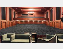 Innenraum eines Theatersaals mit roten Wänden, einer Holzvertäfelung und mehreren Reihen dunkel gepolsterter Sitzplätze, darunter ein Balkon. Im Vordergrund befinden sich grüne Polstersessel und kleine Tische.