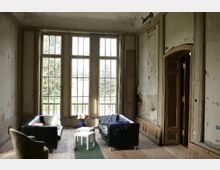 Ein verfallenes Zimmer mit hohen Fenstern, abblätternder Wandfarbe und Sicht auf einen Garten. Im Raum stehen zwei schwarze Sofas, ein Sessel und ein kleiner weißer Tisch auf Holzboden.