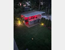 Moderner Garten mit einer kleinen offenen Gartenküche unter einem Flachdach, beleuchtet durch rotes und warmweißes Licht. Im Vordergrund liegt eine Rasenfläche, umgeben von hohen Bäumen und Hecken.