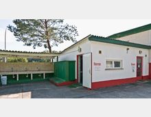 Ein einfacher Sanitärbereich auf einem Campingplatz mit separaten Eingängen für "Herren" und "Damen". Links befindet sich ein überdachter Waschbereich mit mehreren Wasserhähnen an einer gefliesten Wand, während im Hintergrund Bäume sichtbar sind.