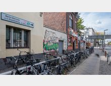 Straßenszene in Berlin mit einem Gehweg, der an einer gelben und roten Hausfassade vorbeiführt, auf denen Graffiti zu sehen sind. Fahrräder sind an einem Geländer abgestellt, daneben ein Wegweiser zur S-Bahn-Station "Berlin-Wannsee/Oranienburg" sowie im Hintergrund eine erhöhte Fußgängerbrücke und weitere Gebäude.