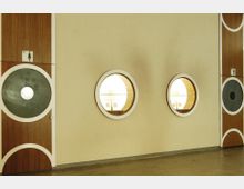 Helle Innenansicht einer Wand mit runden, beleuchteten Fensteröffnungen und zwei Türen mit großen Kreismustern aus Holz und Glas. Auf den Türen sind WC-Schilder für Damen und Herren zu sehen.