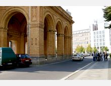 Historisches Backsteingebäude mit dekorativen Bögen und Säulen, flankiert von parkenden Autos. Im Hintergrund befinden sich moderne Gebäude und eine Baumreihe, während rechts eine Gruppe von Fußgängern auf dem Bürgersteig steht.
