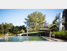 Ein Freibad mit grünlich schimmerndem Wasser, einer großen gewundenen Wasserrutsche und umgeben von Bäumen. Menschen spielen am Beckenrand und genießen die Umgebung bei sonnigem Wetter.