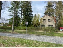 Wohnhaus mit grauer Fassade und rotem Satteldach, umgeben von Bäumen und einem Zaun. Im Vordergrund eine gepflasterte Straße, eine grüne Rasenfläche und ein Verkehrsschild mit der Aufschrift "30 Zone".