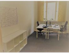 Ein kleiner, heller Büroraum mit zwei weißen Schreibtischen, schwarzen Bürostühlen und einem großen Fenster mit gelben Vorhängen. An der linken Wand hängt ein Jahreskalender, darunter steht ein offenes Regal mit mehreren Fächern.