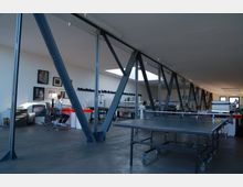 Weitläufiger Arbeitsplatz in einem modernen Raum mit sichtbaren Stahlträgern, weißen Wänden und industriellem Design. Im Vordergrund steht ein Tischtennistisch, im Hintergrund sind Schreibtische, Druckmaschinen und an der Wand aufgehängte Kunstwerke zu sehen.