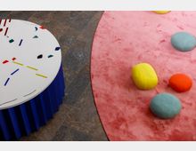 Ein runder Tisch mit weißer Oberfläche, bunten abstrakten Mustern und blauer, plissierter Basis steht auf einem Steinboden. Neben dem Tisch liegt ein großer, pinker Teppich mit bunten, steinförmigen Sitzkissen in Gelb, Blau und Rot.