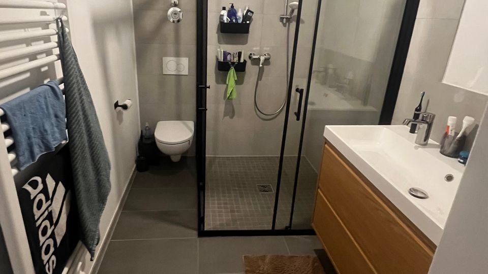 Schmales, modernes Badezimmer mit grauen Boden- und Wandfliesen, bodengleicher Dusche mit schwarzem Rahmen und Glas-Schiebetür. Rechts steht ein Waschtisch mit Holzunterschrank, links ein Handtuchheizkörper; hinten ist ein wandhängendes WC zu sehen.